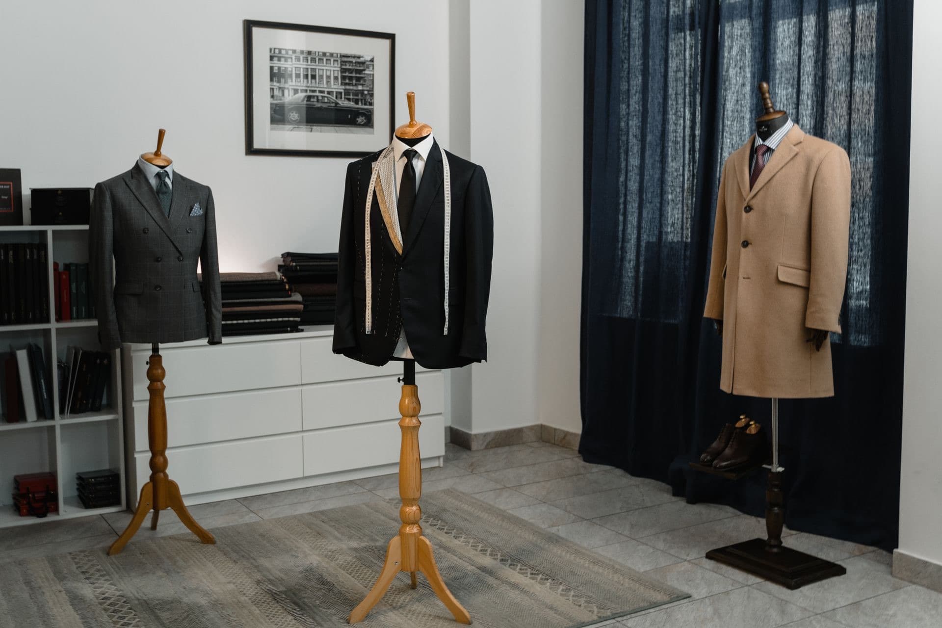 Atelier privado con maniquíes de sastre: saco hilvanado con cinta métrica, traje gris y abrigo camel junto a telas apiladas