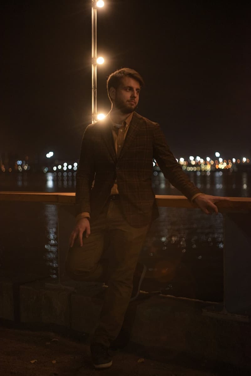 Hombre con blazer de cuadros apoyado en una baranda junto al agua de noche
