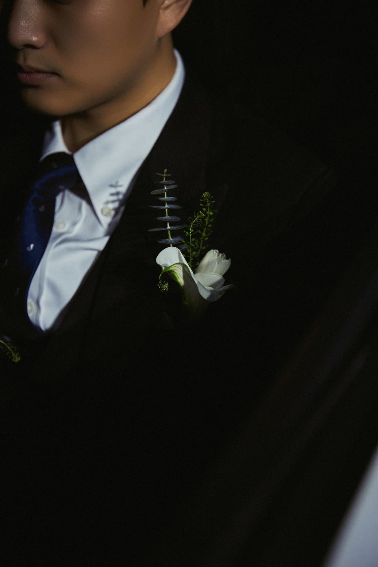Novio con traje oscuro, corbata azul y flor blanca en la solapa, con luz lateral dramática